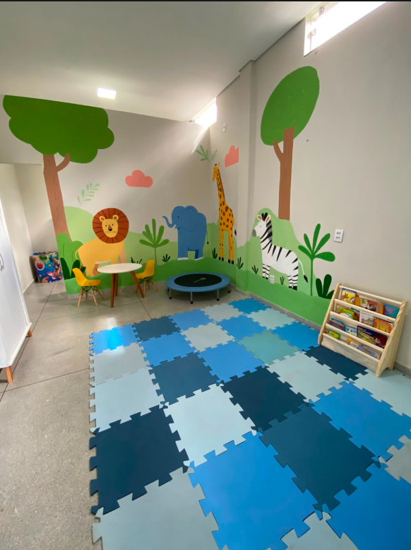 Área infantil com mural de animais, mesa, cadeiras, mini cama elástica e tatame colorido